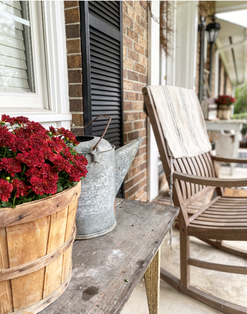 a bushel basket with red mums beside an old watering can on a table near a rocking chair with a grain sack on the back